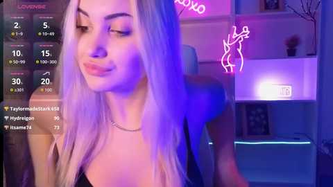 Video of a young, light-skinned woman with long, platinum blonde hair, wearing a black top, in a dimly lit room with neon lights and a TV showing \"The Love Vertigo\" and \"Hydrogen\" videos.