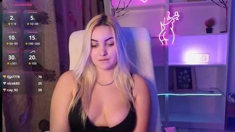 Video of a blonde woman with fair skin, wearing a black bra, sitting in a chair with neon lights and digital stats displayed on a screen.