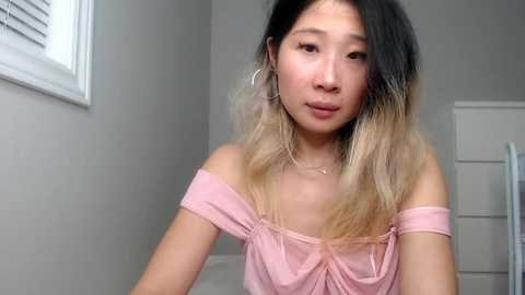 Media: Video of an East Asian woman with long, ombre hair, light skin, and an off-shoulder pink top, standing in a simple, brightly-lit room with white walls and blinds.