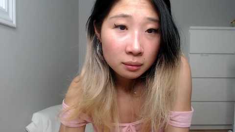 Video of an Asian woman with long, ombre hair, wearing a pink off-shoulder top, in a simple room with white walls and a white dresser. She has a neutral expression.