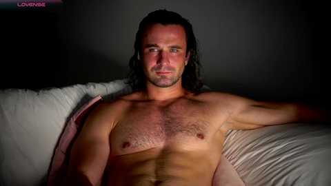 Video of a shirtless man with long, dark hair, lying on a bed, wearing pink shorts, looking serious.