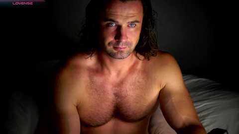 Video of a shirtless, muscular man with long, dark hair, blue eyes, and a subtle smile, seated on a bed with dim lighting, creating a moody, intimate atmosphere.