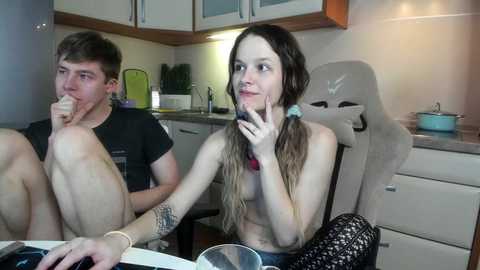 Video of two young, fair-skinned individuals in a modern kitchen, one eating a sandwich, the other with a thoughtful expression.