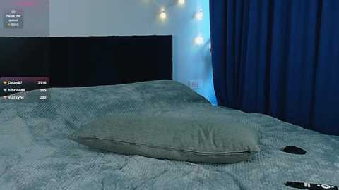 A video of a dimly lit bedroom with a grey pillow on a textured blue bedspread. The background features a dark wooden headboard, a blue curtain, and a glowing string of fairy lights.
