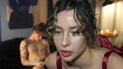 Video of a shirtless man with tattoos, sitting on a black couch, while a woman with wavy, shoulder-length hair and a red dress stands in front of him.