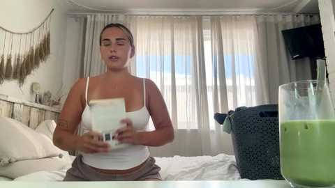 Video of a young woman in a white tank top and beige shorts, holding a green smoothie, standing in a bright, modern bedroom with a large window, white curtains, and a laundry basket.