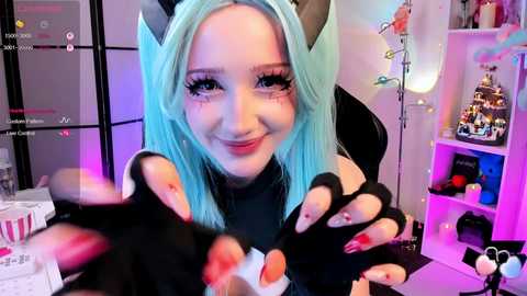 Video of a young woman with light skin and blue hair, wearing a black catsuit with cat ears, posing playfully. Background includes a gaming setup with a monitor, shelves, and toys.