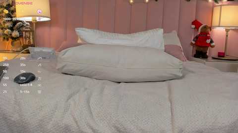 A video of a cozy, well-lit bedroom with a neatly made bed featuring white pillows and a knitted blanket. The background includes a pink upholstered headboard, a nightstand with a lamp, and festive decorations like a Santa Claus figurine.