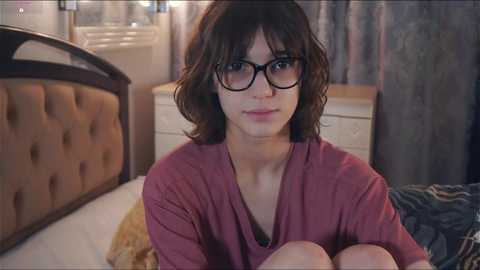 Media: Video of a young woman with shoulder-length brown hair, wearing black-rimmed glasses and a maroon V-neck shirt, sitting on a bed in a dimly lit room with beige walls and a wooden headboard.