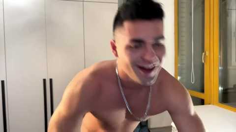 Video of a shirtless, fit young man with short dark hair and a silver chain necklace, laughing with a white tissue covering his mouth, in a modern kitchen with white cabinets and yellow-framed windows.