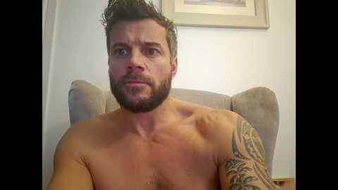 Video of a shirtless, muscular white man with short, dark hair, a beard, and a large tattoo on his right arm. He has a surprised expression, seated on a beige chair against a white wall with framed art.