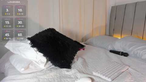 A video of a black cat lying on a white bed with a gray headboard, beside a white keyboard. The background features a dimly lit, beige curtain.