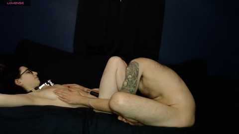 Video of a nude, tattooed woman with glasses and a black collar lying on a black couch, legs bent, hands covering breasts, dark room.