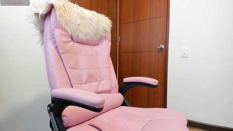 Video of a pink gaming chair with a fluffy white faux fur headrest, black armrests, and a wooden door in a minimalist room.