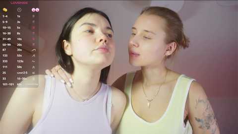Video of two young women, one with short black hair, the other with blonde hair, both wearing tank tops, standing closely with soft lighting.