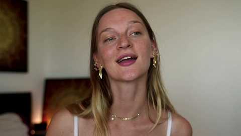 Media: Video of a smiling, fair-skinned, blonde woman with straight hair, wearing a white tank top and gold jewelry, in a dimly lit room with abstract art on beige walls.
