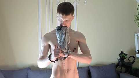 Video of a shirtless, fair-skinned, slim young man with short brown hair, wearing a black leather harness and sucking on a blue denim penis sheath, standing in a dimly lit living room with a beige wall, a purple sofa, and a black cat statue.