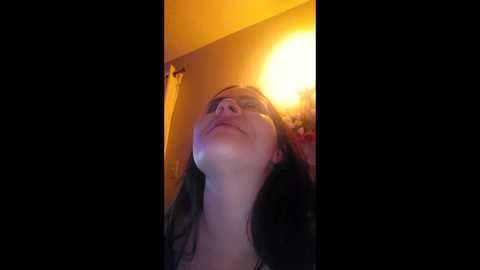 Video of a smiling woman with long dark hair, looking upwards, against a warm, orange-yellow background. She has fair skin and is wearing a black top.