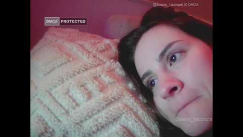 Video of a Caucasian woman with fair skin and brown hair, lying on a beige, textured blanket, under pinkish-red lighting. Her eyes are closed, and her face has a contemplative expression.