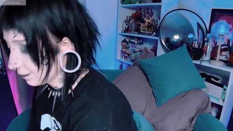 Video of a goth woman with pale skin, black hair, and large silver hoop earrings, wearing a black shirt, sitting on a teal couch, surrounded by a cluttered room with shelves filled with toys, games, and a large round mirror.