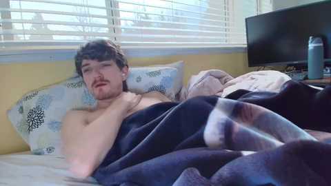 A video of a young, fair-skinned, bearded man with short brown hair, lying on a bed with a blue blanket, looking tired. Background includes a window with white blinds, a TV, and a water bottle.