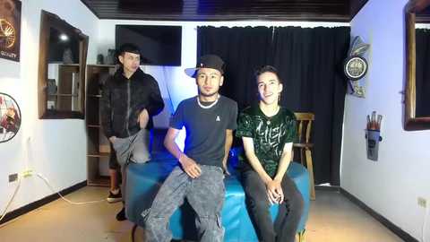 Video of three young men in casual clothes sitting on a blue couch in a dimly lit room with black curtains.