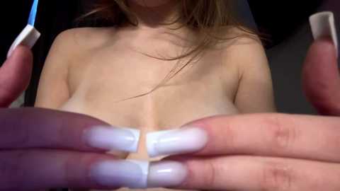 Media: Video of a topless woman with light skin, long brown hair, and large breasts, holding her breasts with manicured hands featuring long, white-painted nails. The background is dark, focusing on her upper body and hands.