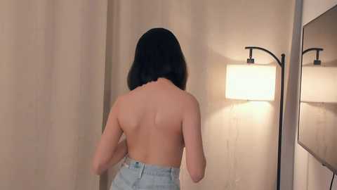 Video of a woman with straight, shoulder-length black hair, standing with her back to the camera, topless, wearing high-waisted light blue jeans. She is in a softly lit room with beige curtains and a floor lamp.