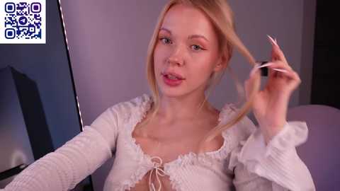 Video of a blonde woman with fair skin, wearing a white lace-trimmed blouse, playing with her hair. Background is a plain wall.