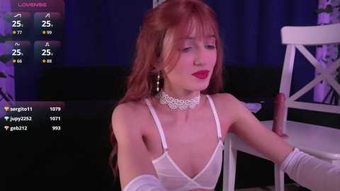 Video of a fair-skinned woman with long red hair, wearing white lingerie and a choker, sitting by a white chair. Background features dark curtains and a blue light.