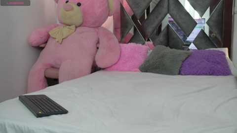 A video of a pink stuffed rabbit with a yellow bowtie leaning against a white wall, with a gray quilted headboard, a white bed, and a black keyboard on the bed.