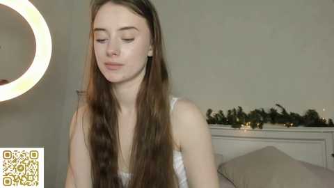 A video of a fair-skinned woman with long brown hair, wearing a white top, standing indoors next to a circular mirror and a garlanded shelf.