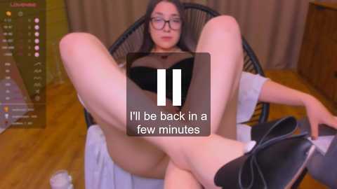 Media: Video of a woman with glasses, black hair, and light skin, wearing a black bra and black high heels, lying on her back in a black wicker chair, legs spread wide, wooden floor, blurred background.