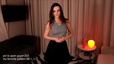 Video of a slender, fair-skinned woman with long, dark hair, wearing a sleeveless gray top and black skirt, standing in a dimly lit living room with beige curtains, a black couch, and a round table with a glowing red lamp.