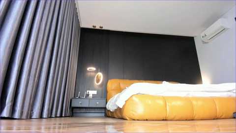 Video of a modern bedroom featuring a yellow tufted leather bed against a black wall, with a grey curtain, bedside table, and circular light fixture.