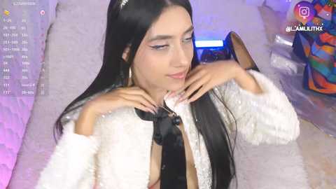 Video of a young woman with long black hair, fair skin, and light makeup, wearing a white fur coat, sitting on a bed. Background includes colorful pillows and a phone screen displaying social media icons.