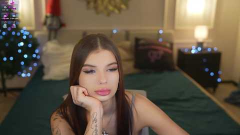 Video of a young woman with long brown hair, wearing makeup, lying on a bed with a Christmas tree and fairy lights in the background.