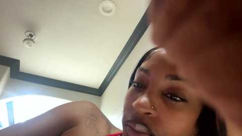 Video of a young African American woman with smooth skin, dark brown eyes, and a small nose piercing, seen from a low angle. She has long, straight black hair and is wearing a red top.