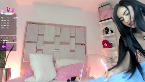 Video of a young woman with long black hair, wearing a black top, standing in a modern bedroom with white walls, a white bed, and pink accents.