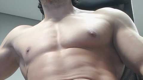 Video of a shirtless man with light skin, muscular physique, and small nipples, standing indoors, with blurred background.