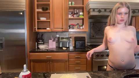 Video of a topless, fair-skinned, blonde woman in a kitchen, wearing only black panties, standing near a granite countertop with cooking utensils and a mixer. Wooden cabinets, stainless steel appliances, and a gas stove are visible in the background.