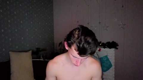 A video of a young, shirtless boy with dark hair and fair skin, sticking out his tongue. The background features a dimly lit room with a gray patterned wallpaper and a white Christmas tree.