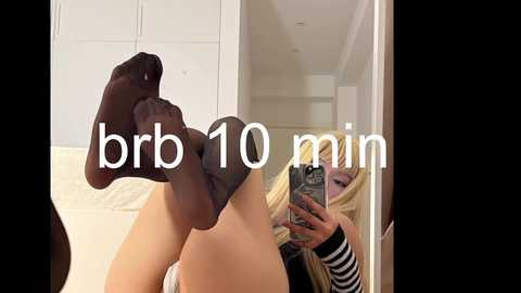 Media: A video of a woman with long blonde hair, wearing black sheer tights, lying on a bed, capturing a selfie in a mirror, with \"bbr 10 min\" text overlay.