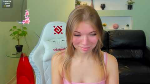 A video of a young woman with light skin and long blonde hair, wearing a pink spaghetti strap top, sitting on a white gaming chair with red accents, in a modern living room with green walls and black leather sofa.