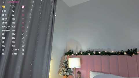 A video of a cozy bedroom with a pink upholstered headboard, green garland, fairy lights, and a white lamp on a nightstand. A gray curtain with sparkly embellishments partially covers the window.