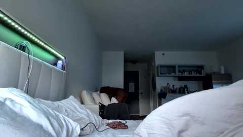 Video of a modern, minimalist bedroom with white walls and bedding. Neon green lights line the headboard, and a person with dark hair, wearing a white shirt and black pants, is sitting on the bed, holding a phone.