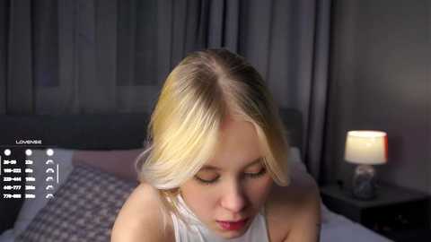 Video of a blonde woman with fair skin, wearing a white sleeveless top, sitting on a bed with gray checkered pillows. The background features a dark gray headboard, curtains, and a nightstand with a lamp.