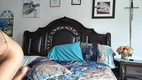 Video of a bedroom with a dark wooden headboard, a turquoise pillow, a floral-patterned bedspread, and a bouquet of flowers on a nightstand, with framed art and a crucifix on the wall.