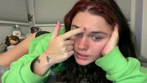 Video of a young woman with long, red-streaked hair, green eyes, and freckles, wearing a green hoodie, making a \"peace\" sign with her hands, in a messy bedroom with a stuffed animal and blinds.