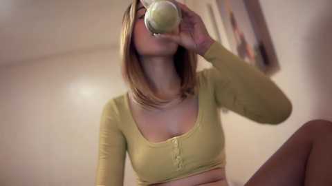 Video of a blonde woman with fair skin, wearing a green crop top, drinking from a green glass, in a dimly lit room with beige walls.